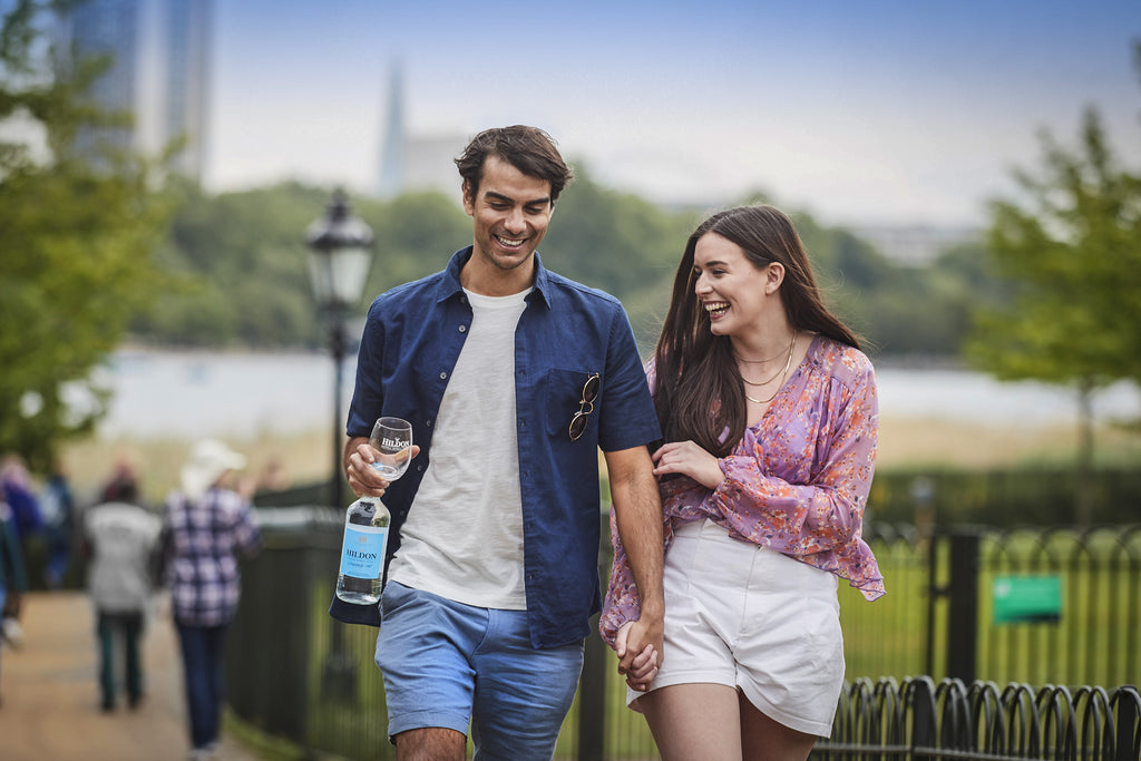 Young couple, walking and laughing in London, holding hands with man carrying a bottle of Hildon still water and a glass.